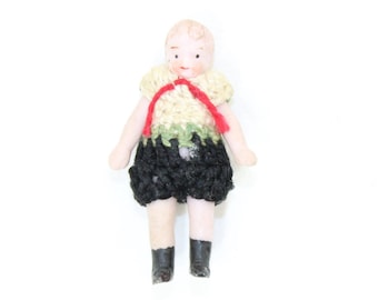Op de afbeelding: Een porseleinen pop met een gehaakt outfit. De pop draagt ​​een geel en groen gehaakt topje met een rood lint en een zwarte gehaakte broek. De pop heeft zwarte schoenen.