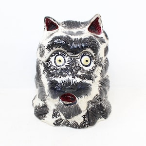 May include: A ceramic dog-shaped container with a distressed, vintage appearance. The dog has black and white fur, with red accents on the ears and mouth. The eyes are yellow with black pupils. The container is likely used for storage or decoration.
