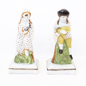 May include: A pair of vintage porcelain figurines depicting a girl and a boy. The girl is wearing a white dress with brown polka dots and a brown hooded cloak. She is holding a small bouquet of flowers. The boy is wearing a brown jacket with white polka dots, yellow pants, and a black hat. He is holding a sheaf of wheat. Both figurines are sitting on green bases.
