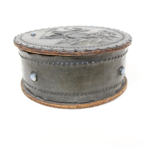 May include: A round, gray leather box with a raised design on the lid. The box has a wooden rim and is decorated with three blue gemstones.
