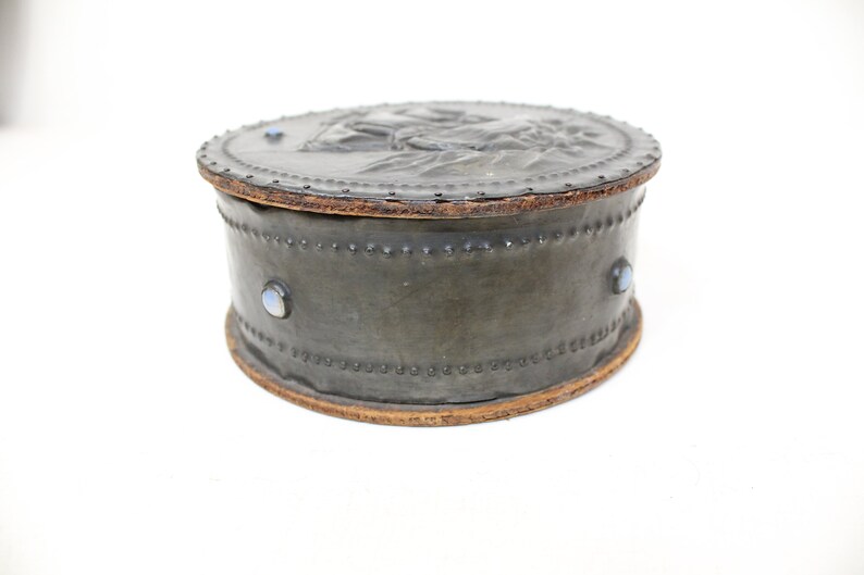 May include: A round, gray leather box with a raised design on the lid. The box has a wooden rim and is decorated with three blue gemstones.