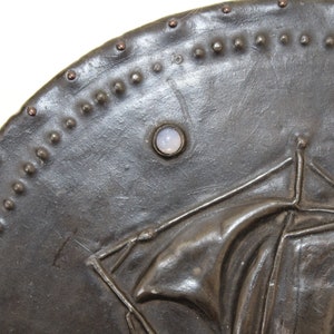 May include: A dark gray metal shield with a raised design of a figure holding a staff. The shield is decorated with a row of small metal studs around the edge and a single white stone set in the center.