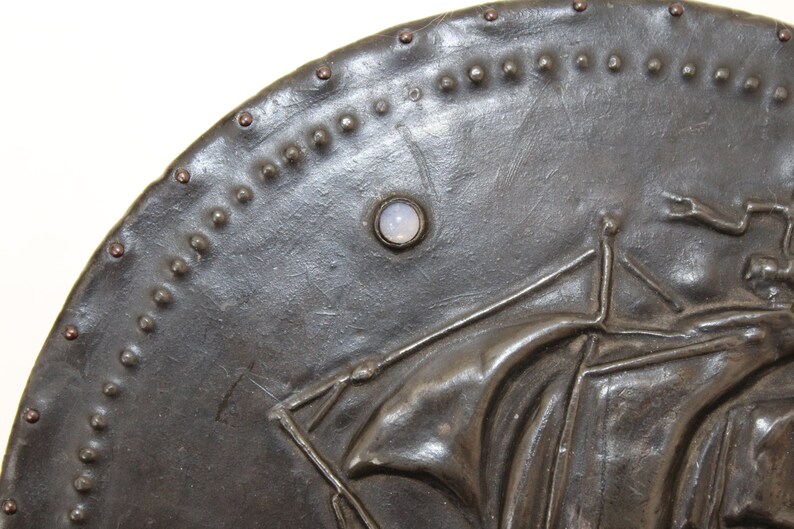 May include: A dark gray metal shield with a raised design of a figure holding a staff. The shield is decorated with a row of small metal studs around the edge and a single white stone set in the center.