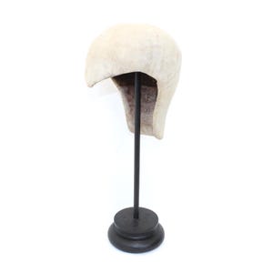 May include: A cream-colored, vintage-style hat, possibly a helmet, displayed on a black metal stand. The hat has a rounded top and a curved brim, with a darker interior. The stand has a circular base.