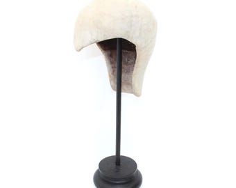 May include: A cream-coloured, vintage-style hat, possibly a helmet, displayed on a black metal stand. The hat has a rounded top and a curved brim, with a darker interior. The stand has a circular base.