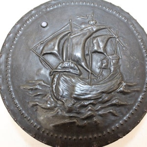 May include: A round, black metal shield with a raised design of a ship sailing on the waves. The shield is decorated with a row of small metal studs around the edge.