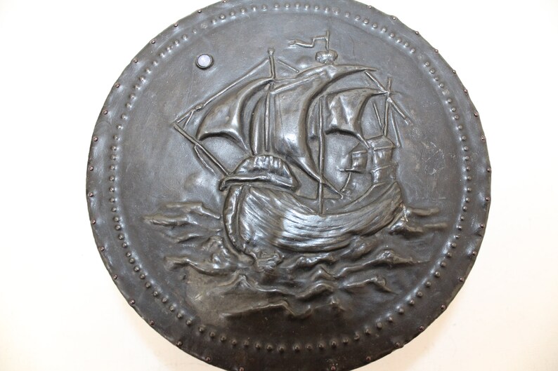 May include: A round, black metal shield with a raised design of a ship sailing on the waves. The shield is decorated with a row of small metal studs around the edge.