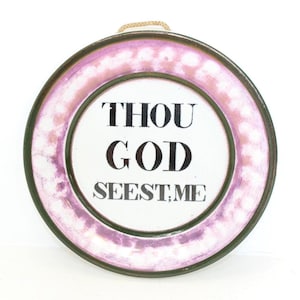 May include: Circular decorative plate featuring the words "THOU GOD SEEST ME" in black. The plate has a white centre, a pink and white dotted border, and a dark green rim. A woven cord is attached for hanging.