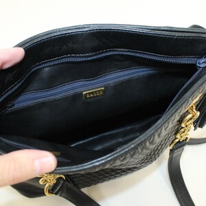 May include: Black quilted leather handbag with gold-tone chain and tassel details. The interior features a navy blue zipper and a gold "BALLY" logo. The bag has a shoulder strap and is open, revealing the inside.