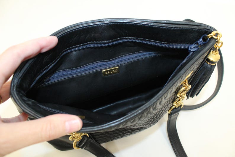 May include: Black quilted leather handbag with gold-tone chain and tassel details. The interior features a navy blue zipper and a gold "BALLY" logo. The bag has a shoulder strap and is open, revealing the inside.