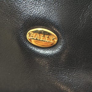 May include: Close-up of a black leather item with a gold oval emblem. The emblem features the brand name "BALLY" in gold lettering. The leather has a textured appearance.