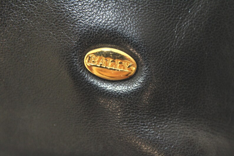 May include: Close-up of a black leather item with a gold oval emblem. The emblem features the brand name "BALLY" in gold lettering. The leather has a textured appearance.