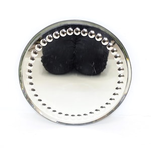 May include: A circular wall mirror featuring a reflective glass surface framed by a border of small, polished silver-toned metallic spheres. The mirror reflects a dark, rounded object in its center, creating a clear contrast against the metallic beads.