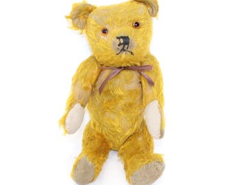 May include: A vintage, golden-yellow teddy bear with a stitched black nose and brown eyes. The bear has a brown ribbon tied around its neck and off-white paw pads. The plush toy is standing upright against a white background.