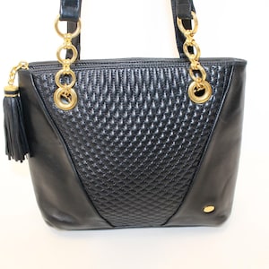 May include: Black leather tote bag with gold-tone chain handles and accents. The center panel features a quilted diamond pattern. A black tassel hangs from the zipper pull. A small gold-tone accent is on the lower right side.