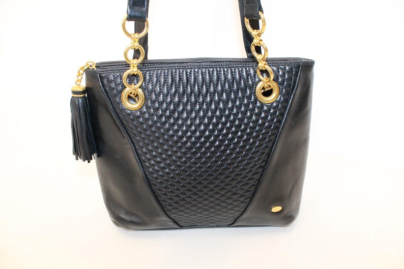 May include: Black leather tote bag with gold-tone chain handles and accents. The center panel features a quilted diamond pattern. A black tassel hangs from the zipper pull. A small gold-tone accent is on the lower right side.