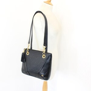 May include: Black leather tote bag with gold-tone chain accents and a tassel. The bag features a textured front panel and long shoulder straps. The bag is displayed on a mannequin.