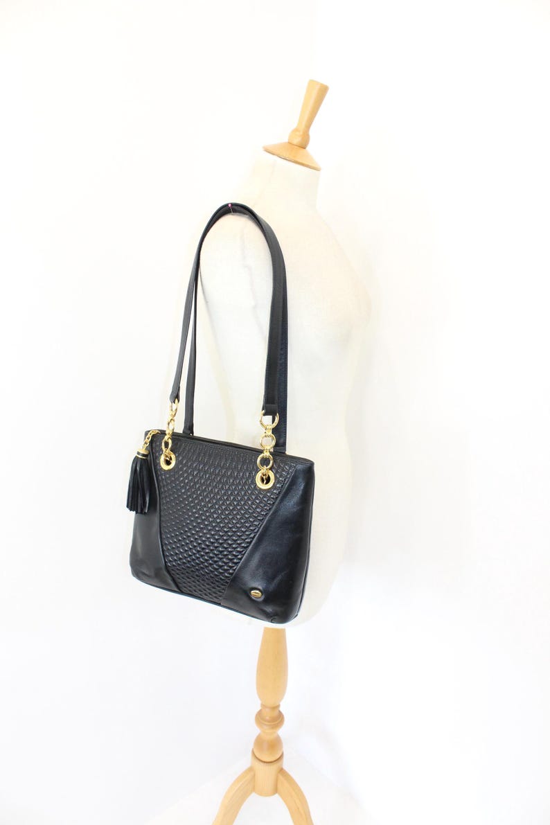 May include: Black leather tote bag with gold-tone chain accents and a tassel. The bag features a textured front panel and long shoulder straps. The bag is displayed on a mannequin.