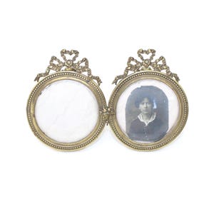 Puede incluir: Un marco de fotos ovalado doble vintage en metal dorado. El marco es articulado y presenta un retrato en un lado. Cada óvalo está rematado con un lazo decorativo. El marco está sobre un fondo blanco.