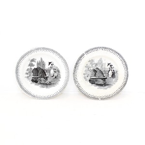 May include: A pair of white ceramic plates, each with a black and white rural scene. The plates have a detailed illustration of figures near a structure, with a decorative border. These plates are likely vintage or antique.