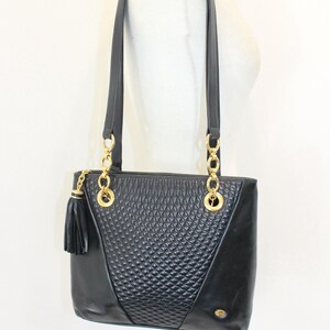 May include: Black leather tote bag with gold-tone chain accents and a tassel. The bag features a quilted diamond pattern on the front and a rectangular shape with two top handles.