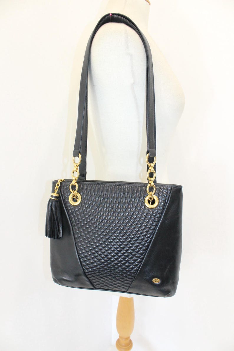 May include: Black leather tote bag with gold-tone chain accents and a tassel. The bag features a quilted diamond pattern on the front and a rectangular shape with two top handles.