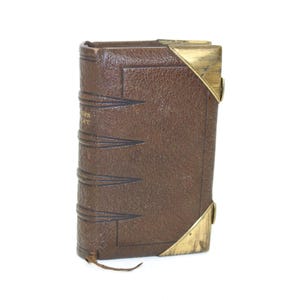 Peut inclure: Un petit livre rectangulaire relié en cuir marron à la texture grainée. Le dos présente des bandes horizontales en relief et des lettres dorées estompées. La couverture possède des coins en laiton en haut et en bas à droite, avec un lien en cuir.