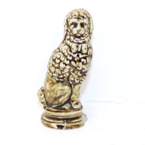May include: A ceramic dog figurine in shades of brown and beige. The dog is seated, with a detailed, textured coat and a round base. The figurine has a glossy finish and is set against a white background.
