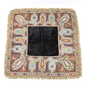 May include: Square brown textile with a black center and paisley pattern border. The border features colorful paisley designs in shades of blue, orange, and green, with a fringed edge. The textile is likely a decorative item.