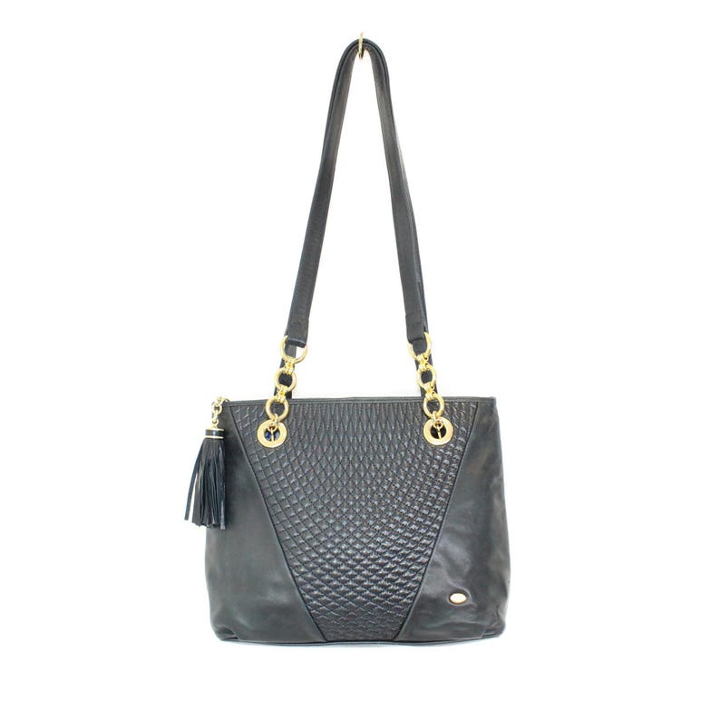 May include: Black leather tote bag with gold-tone chain straps and accents. The bag features a quilted diamond pattern on the front and a black tassel. The bag has a rectangular shape and long shoulder straps.