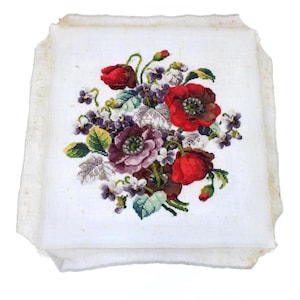 May include: A square, off-white fabric with a detailed cross-stitch floral design. The bouquet features vibrant red poppies, purple violets, and green leaves. The edges of the fabric are slightly frayed.
