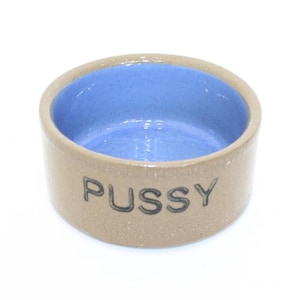 Puede incluir: Un cuenco de cerámica pequeño y redondo con exterior marrón claro e interior azul. La palabra "PUSSY" está impresa en negro en el lateral del cuenco. El cuenco es probablemente para una mascota.