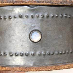 May include: Close-up of a metal container with a circular, translucent, light blue glass insert. The container has a brown leather rim and a row of small metal rivets around the top and bottom.