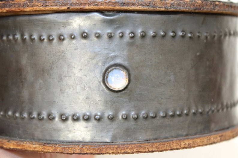 May include: Close-up of a metal container with a circular, translucent, light blue glass insert. The container has a brown leather rim and a row of small metal rivets around the top and bottom.