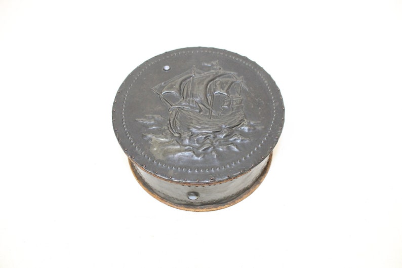 May include: A round, black, wooden box with a raised, embossed design of a ship on the lid. The box has a brown wooden base.