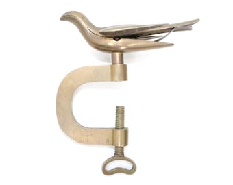 May include: A brass-colored bird-shaped clamp, featuring a dove design. The clamp has a C-shaped body with a threaded screw and a handle for adjustment. The bird's wings are slightly spread, and the clamp is set against a white background.