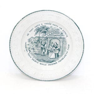 May include: White ceramic plate with a teal border and a detailed illustration. The illustration shows a blacksmith and other figures near a structure, with text around the rim and center. The text reads: "HE THAT HATH A TRADE HATH AN ESTATE. INDUSTRY PAYS DEBTS WHILE DESPAIR INCREASETH THEM."