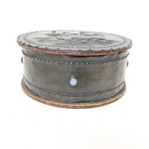 May include: A round, gray leather box with a decorative design on the lid. The box is trimmed with a brown leather band and has small, white stones around the edge.
