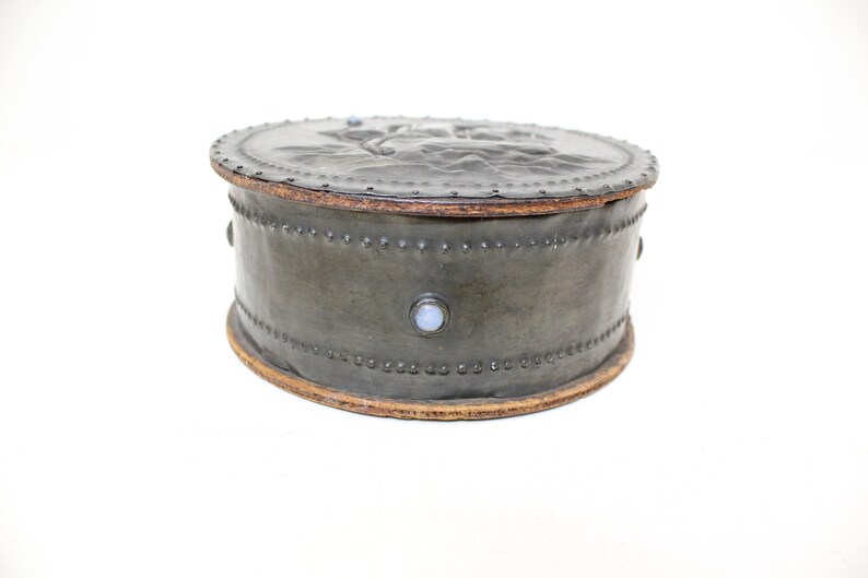 May include: A round, gray leather box with a decorative design on the lid. The box is trimmed with a brown leather band and has small, white stones around the edge.