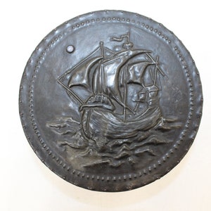 May include: A round, metal wall plaque with a raised design of a ship sailing on the waves. The plaque has a dark gray finish and is decorated with a border of small, raised dots.