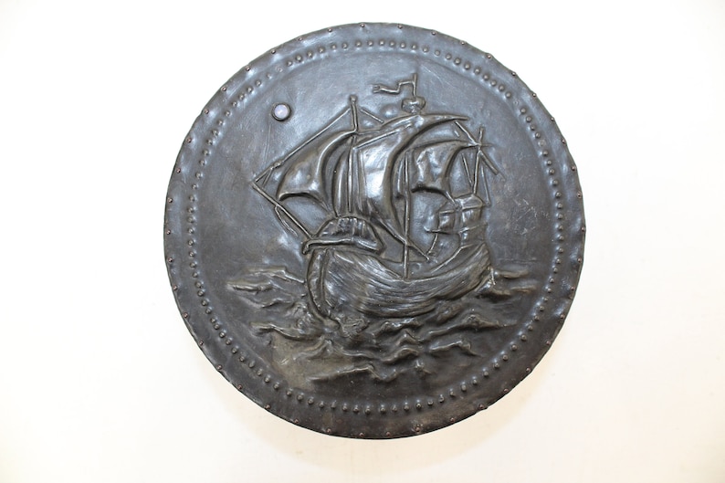 May include: A round, metal wall plaque with a raised design of a ship sailing on the waves. The plaque has a dark gray finish and is decorated with a border of small, raised dots.