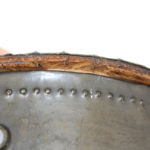 May include: Close-up of the top edge of a metal container with a wooden rim. The metal is silver and has a series of small, evenly spaced rivets around the edge. The wooden rim is brown and has a slightly rough texture.