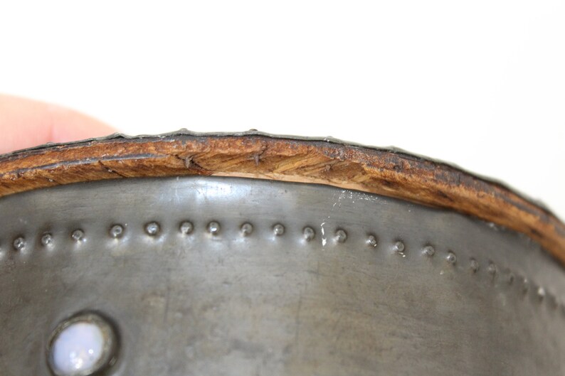 May include: Close-up of the top edge of a metal container with a wooden rim. The metal is silver and has a series of small, evenly spaced rivets around the edge. The wooden rim is brown and has a slightly rough texture.
