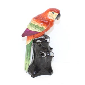 May include: A ceramic parrot ornament perched on a black tree stump. The parrot has red, orange, green, and purple feathers, with a yellow and black beak. The tree stump has several holes with white accents, creating a decorative ornament.
