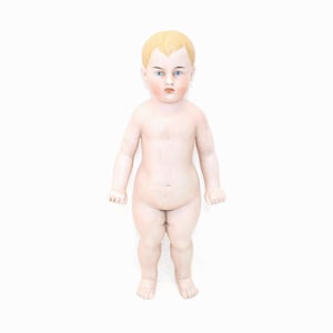 May include: A porcelain doll of a baby with blonde hair and blue eyes. The doll has a pale complexion and is nude. The doll is standing upright with its arms at its sides. The doll's face has a slight blush.