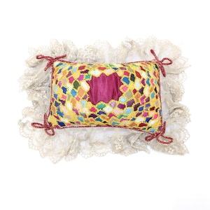 May include: A decorative pillow with a patchwork design in various colors, including yellow, pink, blue, and green. The pillow has a central magenta-colored section and is trimmed with gold thread. It is surrounded by a cream-colored lace trim and tied with red bows.