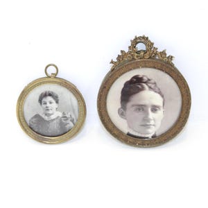 May include: Two antique portrait miniatures. One is round with a simple gold frame and a small loop at the top. The other is oval with an ornate gold frame. Both feature black and white portraits of women.
