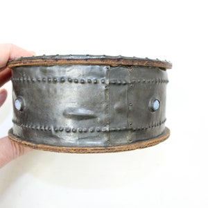 May include: A round, silver metal box with a wooden top and bottom. The box has a decorative pattern of small metal studs around the sides. There are two small, round, white accents on the front of the box.
