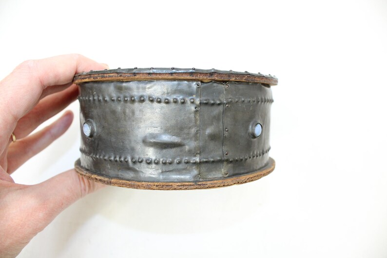 May include: A round, silver metal box with a wooden top and bottom. The box has a decorative pattern of small metal studs around the sides. There are two small, round, white accents on the front of the box.