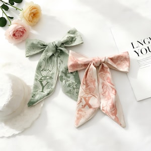 Op de afbeelding: Twee zijden haarsjaals in de vorm van een strik. Eén is lichtgroen met een bloemen- en letter "Y"-ontwerp. De andere is blush roze met een bloemen- en letter "X"-ontwerp. De sjaals liggen op een wit oppervlak met bloemen.